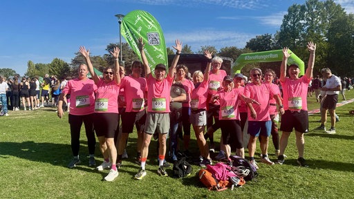 Eine Gruppe Menschen mit pinken T-Shirts steht jubelnd auf einer grünen Wiese unter blauem Himmel.