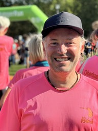 Ein Mann mit schwarzer Kappe und in pinkem T-Shirt lächelt in die Kamera. Hinter ihm stehen viele Menschen einer Bühne zugewandt.