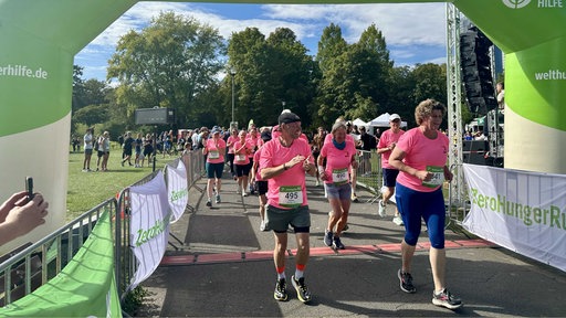 Viele Menschen mit pinken-Shirts auf denen Startnummern klebenund Sporthosen laufen einen grünen Zieltunnel ein.