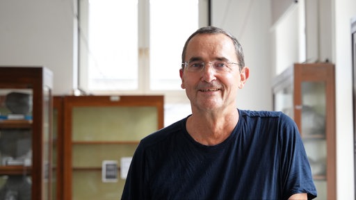 Ein mittelalter Mann mit Brille und blauem T-Shirt lächelt in die Kamera, im Hintergrund alte Vitrinen