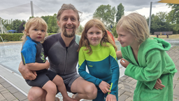 Ein Vater mit drei Kinder hockt in einem Freibad, vor einem Schwimmbecken vor der Kamera. 