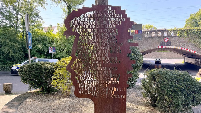 Ein Mahnmal aus rostigem Stahl in Form eines Gesichtprofiles mit eingelassenem Text steht in einem mit Büschen umringten Kiesbett.