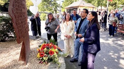 Vor einer Gedenkstele aus Stahl liegt ein bunter Blumenkranz, davor stehen Personen in förmlicher Kleidung und schauen hinauf.