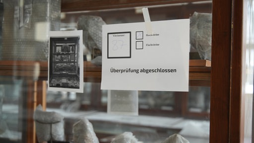 Eine Vitrine im Goldfußmuseum. Die Gegenstände in ihr sind noch verpackt. Ein Zettel hängt am Glas. "Überprüfung abgeschlossen."