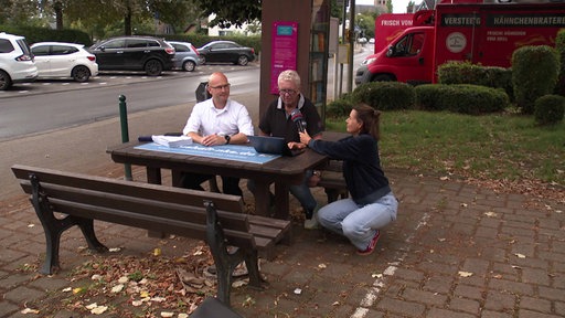 Zwei Männer sitzen auf einer Bank, vor ihnen liegt ein Papierstapel. Eine Reporterin hockt mit einem Mikrofon neben ihnen. Sie befinden sich neben einer Straße mit parkenden Autos.
