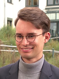 Ein junger Mann mir grauem Anzug und Brille. Er hat brünette Haare die zur Seite liegen. 