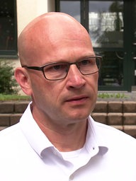 Ein Mann mit Glatze, einem weißen Hemd und einer schwarzen Brille. 
