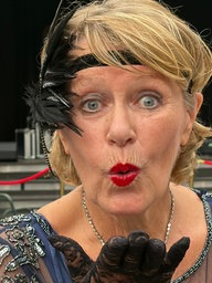 Eine Frau mit blonden kurzen Haaren hat ein schwarzes Haarband im Stil der 20-er Jahre an. Sie trägt roten Lippenstift und ein schwarzes Kleid und Handschuhe. Sie schaut in die Kamera und wirft einen Luftkuss zu. 