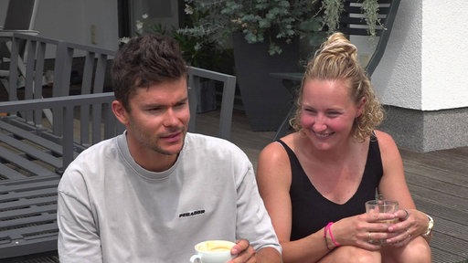 Jan Stratmann sitzt mit seiner Frau auf einer Terrasse. Hinter ihnen stehen Gartenmöbel und Pflanzen. Beide halten eine Kaffeetasse in der Hand.