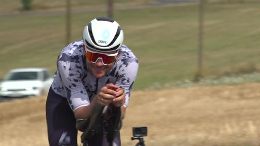 Ein Rennradfahrer sitzt dynamisch auf dem Rennrad. Die Landschaft im Hintergrund ist verschwommen. Er trägt Brille, Trikot und Helm
