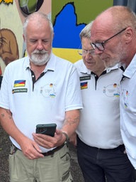 Vier Männer stehen vor einem LKW. Sie schauen auf ein Handy, auf das einer von ihnen zeigt. Sie tragen weiße Hemden.