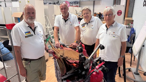 Vier Männer stehen hinter einem Einkaufswagen mit verschiedenen Gegenständen. Sie tragen weiße Hemden und stehen in einem Raum mit Gebrauchsutensilien.