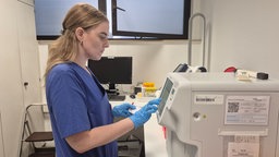 Eine junge Krankenpflegerin steht in blauer Arbeitskleidung im Labor und schwitzt.