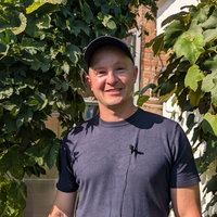 Ein Mann mit einem dunkelblauen T-Shirt und einer dunkelblauen Schirmkappe lächelt in die Kamera. Hinter ihm sieht man eine grüne Pflanze. 
