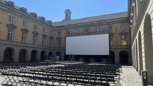 Im Arkadenhof der Universität Bonn stehen schwarze Klappstühle aufgereiht vor einer großen Leinwand.