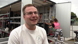 Mann Mitte 30 mit weißem Shirt und kurzen Haaren, im Hintergrund ein LKW und tragende Menschen