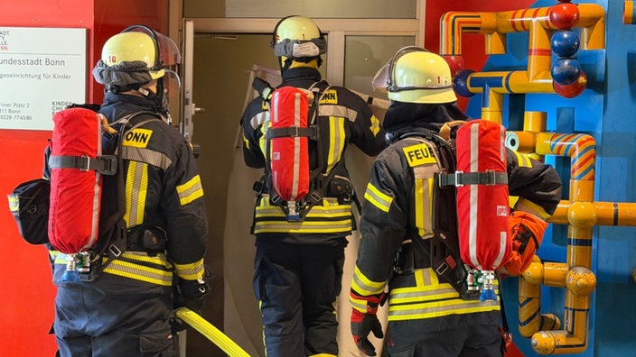 Drei Feuerwehrleute tragen Helme und Schutzkleidung. Sie betreten ein Gebäude. Der Feuerwehrmann in der Mitte hat die Tür aus den Angeln gehoben. In dem Raum sieht man viel Rauch. 