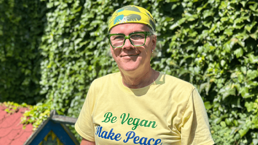 Eine Frau mit einer gruenen Brille, gelben T-shirt und einem bunten Tuch auf dem Kopf vor einer bepflanzten Wand.