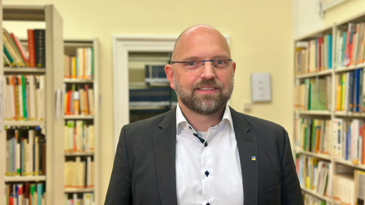 Ein Mann mit Brille und Anzug in einem Raum voller Bücherregale.