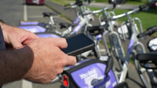 Zwei Hände halten ein Smartphone. Im Hintergrund stehen Leihfahrräder.