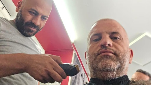 Ein Mann mit kurz geschnittenen Haaren im Friseursalon. Neben ihm steht der Friseur, der seine Haare schneidet.