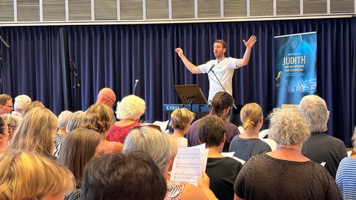 der Chorleiter steht während einer Chorprobe erhöht auf einer Bühne und dirigiert rund 150 Sänger