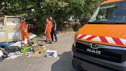 Links im Bild stehen Männer in orangener Arbeitskleidung vor einem Berg Müll, rechts steht ein orangenes Fahrzeug.