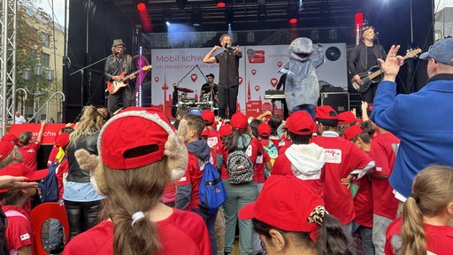 Eine Band spielt auf einer Bühne. Im Publikum tragen die Zuschauer rote Kleidung.