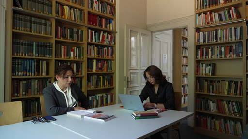 Zwei Frauen sitzen an einem Tisch und lernen, im Hintergrund stehen Bücherregale