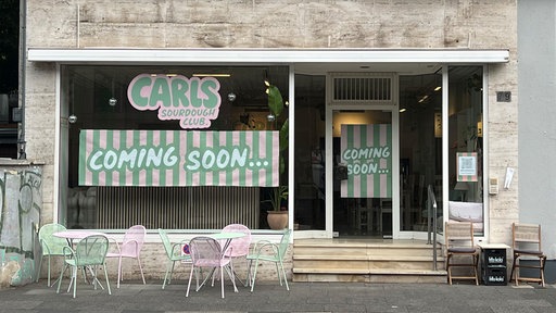 pinke und grüne Stühle stehen vor einer Fensterfront und Glastür, an der Glaswand ist ein Plakat mit Beschriftung "Coming Soon" angebracht