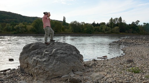 Ein Mann mit rosa Oberteil steht zum fotografieren auf einem großen Stein am Rhein