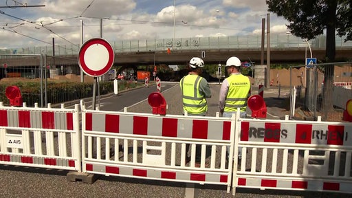 Ein rot-weißer Absperrzaun, daneben zwei Bauarbeiter in gelben Warnwesten, im Hintergrund die Bonner Vorlandbrücke.