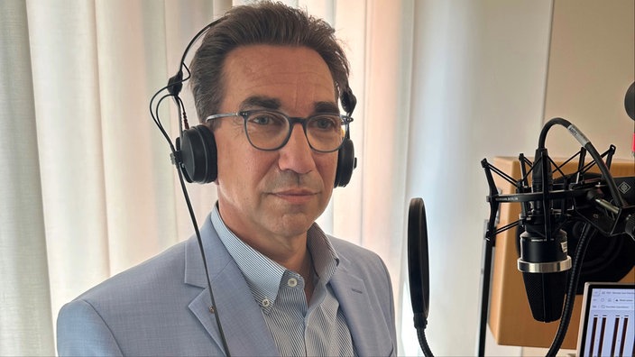 Ein Mann mit Brille und dunklen Haaren trägt Kopfhörer und steht vor einem Mikrofon