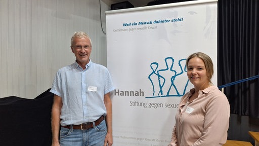 Ein Mann und eine Frau stehen vor einem Banner auf dem "Hannah. Stifung gegen sexuelle Gewalt steht".