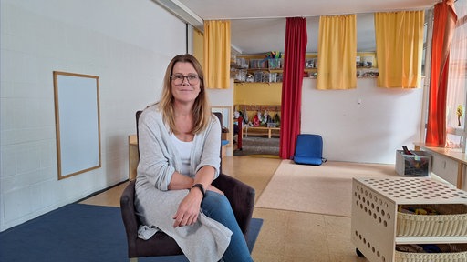 Eine Frau sitzt auf einem Sessel in der Mitte eines Raums. Im hINtergrund bunte Vorhänge.