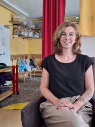 Eine Frau sitzt auf einem Stuhl. Sie trägt ein schwarzes Shirt, eine Brille und lächelt.