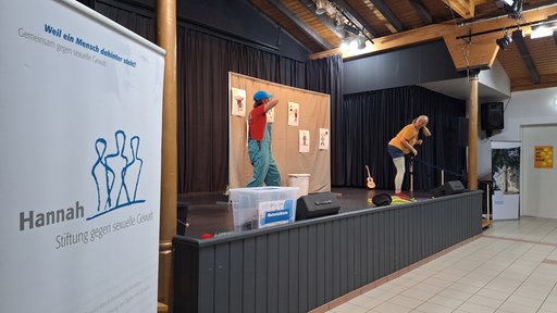Zwei Menschen stehen verkleidet auf einer Bühne. Links im Bild ein Banner mit der Aufschrift: "Stiftung gegen sexuelle Gewalt"