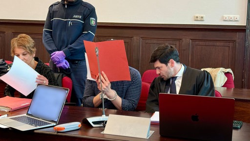Ein Mann sitzt verdeckt mit einer Mappe sein Gesicht. Er sitzt in einem Gerichtssaal. Rechts neben ihm sitzt sein ein weiterer Mann in Anzug. Links sitzt eine Frau, die sich ein Blatt anschaut. Im Hintergrund steht ein Polizist.