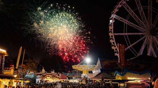 Viele Menschen tummeln sich bei Dunkelheit zwischen den Fahrgeschäften, am Himmel blitzt ein Feuerwerk auf.