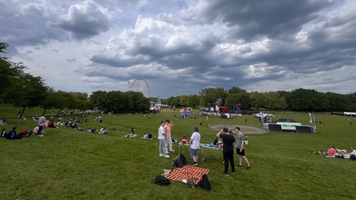 Vereinzelte Besucher stehen in der Bonner Rheinaue. Andere sitzen auf Picknick-Decken. Im Hintergrund ist ein Riesenrad zu sehen. 