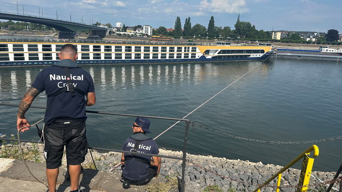 Zwei Männer stehen an dem Ufer und ziehen an eine Seil, das mit einem Hotelschiff verbunden ist, ein Abschleppschiff steht im Hintergrund.