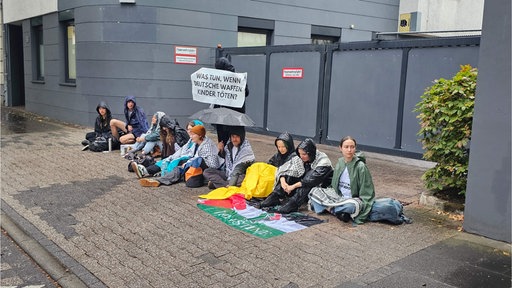 Eine Reihe sitzender Menschen vor einer Einfahrt. Sie tragen Regenjacken, einen Schirm und ein Plakat.