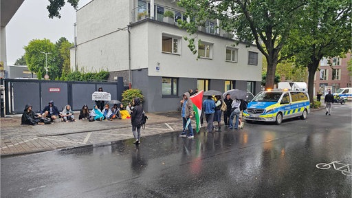 Eine Reihe sitzender Menschen vor einer Einfahrt. Davor eine stehende Gruppe mit Regenschirmen und Fahnen. Daneben ein Polizeiwagen auf der Straße.