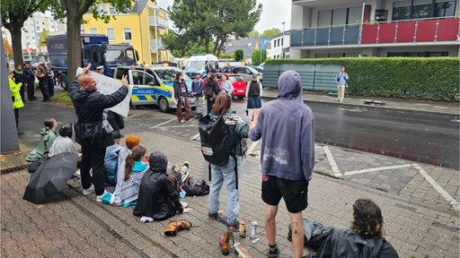 Sitzende und stehende Menschen auf dem Bürgersteig. Weitere Demonstranten und Polizisten auf der Straße.