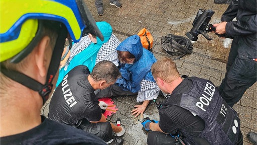 Eine Demonstrantin sitzt auf dem Boden. Ihre Hand klebt am Boden. Zwei Polizisten hocken über der Hand.