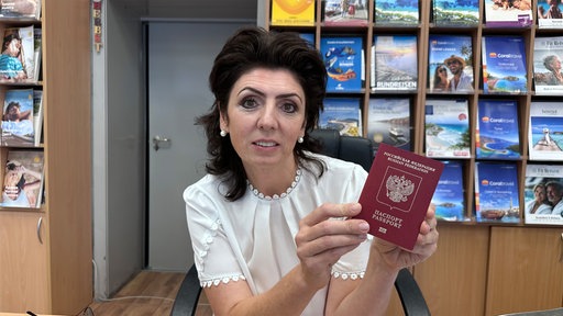 Eine Frau mit dunklen Haaren sitzt auf einem Stuhl. In ihren Händen hält sie einen russischen Pass. Im Hintergrund sind Reisebroschüren.
