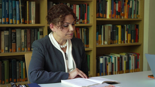 Eine Frau sitzt an einem Tisch vor einem Bücherregal und liest in einem Buch