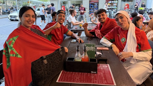 Vier Marokko-Fans sitzen in Trikots im Café und lächeln in die Kamera. 