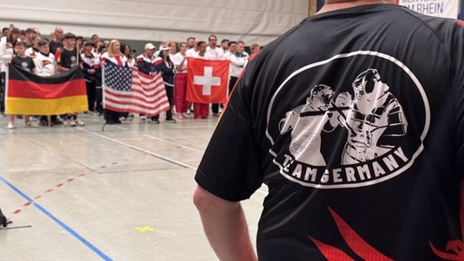 Man sieht von Hinten einen Mann mit einem Tshirt mit der Aufschrift: "Team Germany". Im Hintergrund Personen die die Flaggen der USA, von Deutschland und der Schweiz hochhalten.