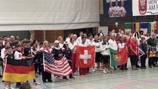 Eine Gruppe von Menschen stehen in einer Sporthalle und halten ihre Nationalflaggen hoch 